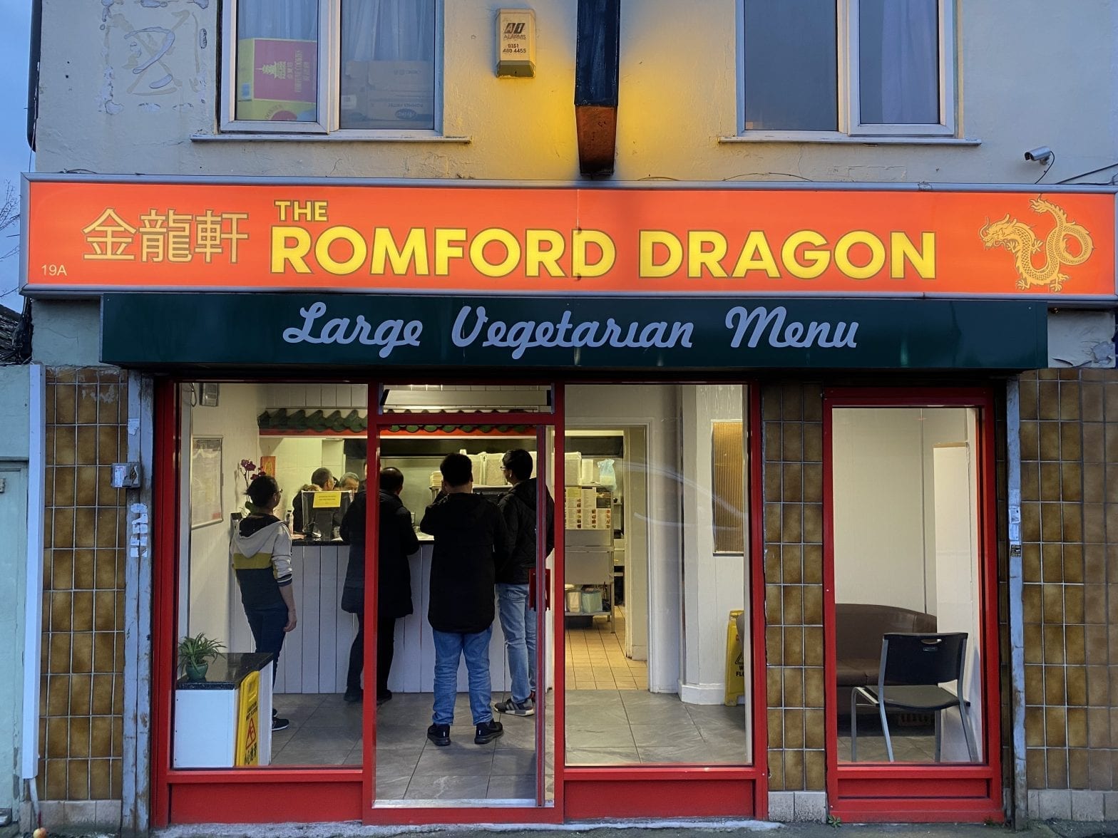 A shopfront with red doors and wide windows. The sign on top in red and gold states the name 'The Romford Dragon' with the address '19A The Parade, Colchester Road, Romford, RM3 0AQ' and finally the phone number '01708 3381818'. Inside the shop looks busy with a cue of people waiting as a woman behind the counter takes orders.